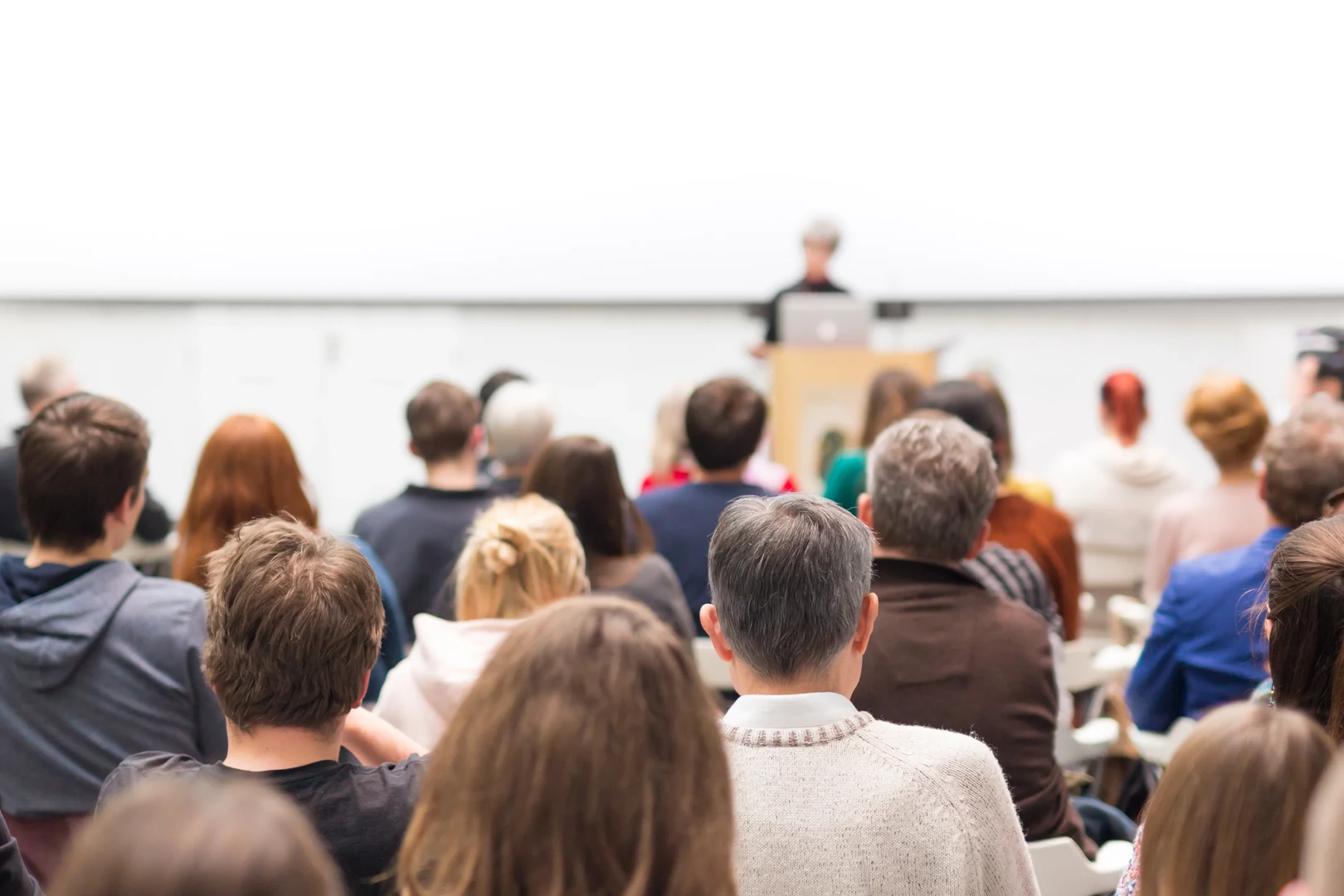 Eine Vortragszene: ein Publikum von hinten, in einiger Entfernung verschwommen eine*n Redner*in an einem Rednerpult.