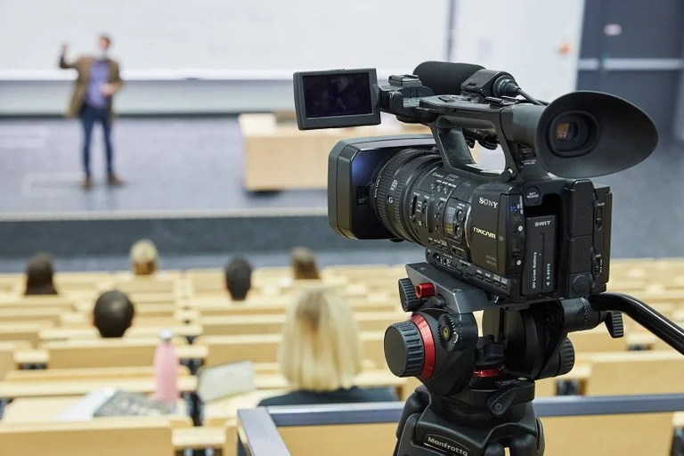 Blick von hinten in einen Vorlesungssaal, im Hintergrund eine vortragende Person (unscharf), vorne eine Videokamera, die auf die vortragende Person gerichtet ist.