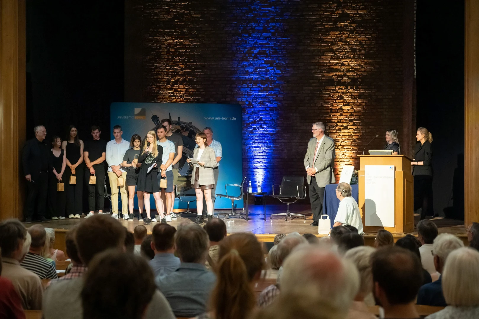 Eine Wissenschaftlerin und ein Wissenschaftler arbeiten hinter einer Glasfassade und mischen Chemikalien mit Großgeräten.