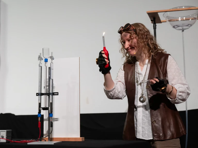 Die Menschheit (Sophia Paul) macht eine Knallgasprobe und zeigt damit, dass bei der Elektrolyse Wasserstoffgas entsteht.