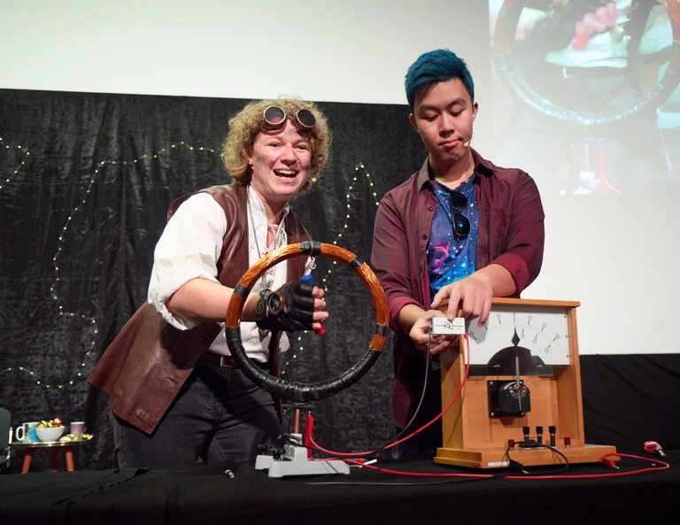 Die Menschheit (Jana Heysel) und das Universum (Tien-Son Nguyen) demonstrieren, wie mit einem Generator Strom erzeugt und eine LED zum Leuchten gebracht werden kann.