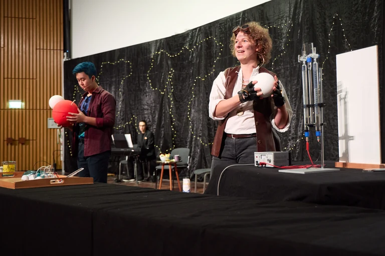 Die Menschheit (Jana Heysel) und das Universum (Tien-Son Nguyen) veranschaulichen, wie bei der Elektrolyse Wasserstoffgas entsteht.