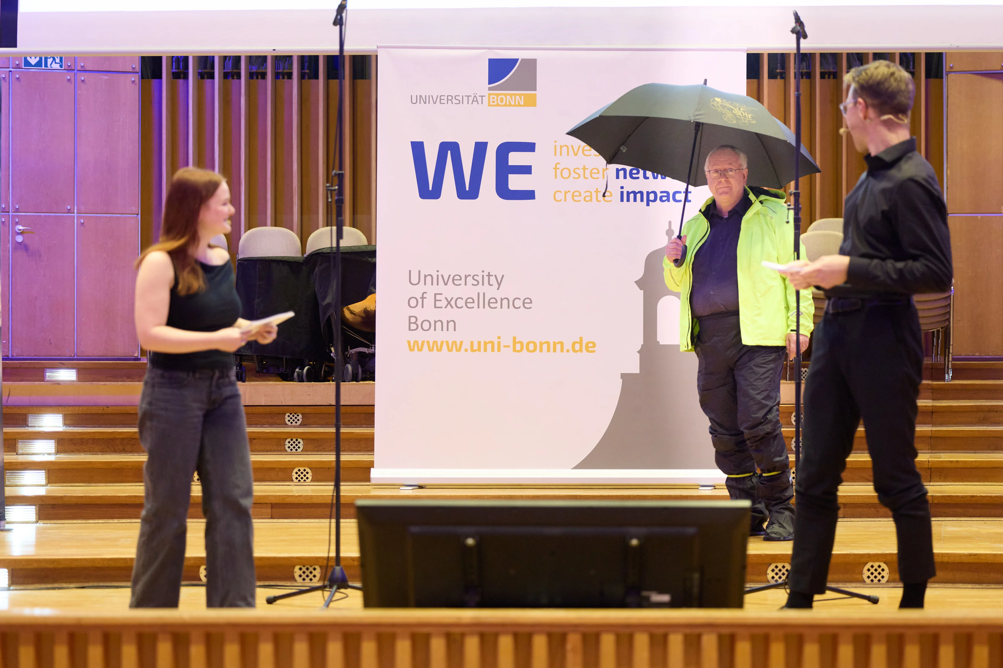 Rektor Prof. Dr. Dr. h.c. Michael Hoch betritt die Bühne – sichtlich auf Extremwetter vorbereitet. Im Vordergrund: Moderator*innen Alea Sonntag und Frederik Schumacher vom Campusradio Bonn FM.