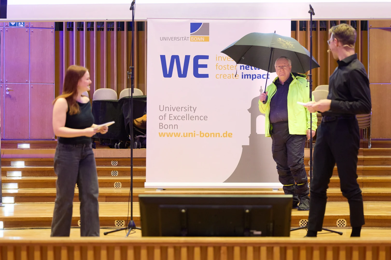Rektor Prof. Dr. Dr. h.c. Michael Hoch betritt die Bühne – sichtlich auf Extremwetter vorbereitet. Im Vordergrund: Moderator*innen Alea Sonntag und Frederik Schumacher vom Campusradio Bonn FM.