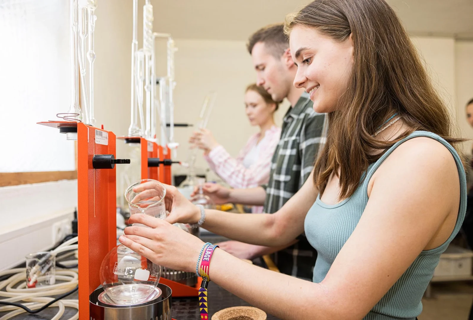 Eine Wissenschaftlerin und ein Wissenschaftler arbeiten hinter einer Glasfassade und mischen Chemikalien mit Großgeräten.