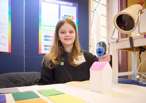 Ein Mädchen sitzt an einem Tisch vor einer Wand. Auf der Wand sind selbsterstellte Poster aufgehängt. Auf dem Tisch liegen bunte Papierstücke.