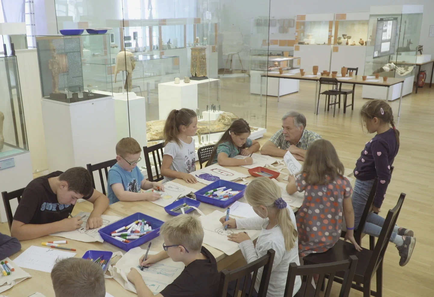 Eine Gruppe Kinder sitzt an einem Tisch im ägyptischen Museum, malt Ausmalbilder und hört einem Dozenten zu.