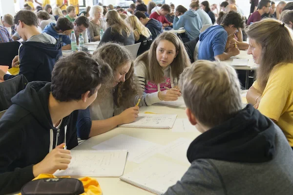 Eine Gruppe Schüler*innen sitzt am Tisch und  diskutiert über Aufgaben auf Arbeitsblättern in einem Raum voller Schüler*innen.
