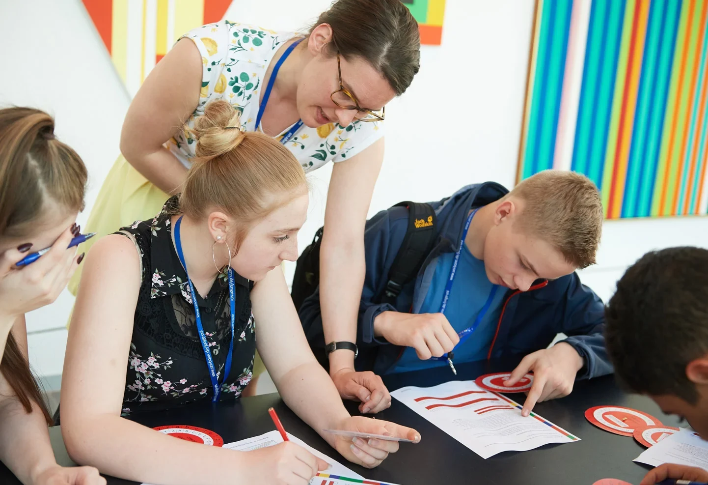 Schüler*innen sitzen an einem Tisch im Arithmeum und lösen Arbeitsblätter, eine Frau beugt sich über sie und hilft ihnen.