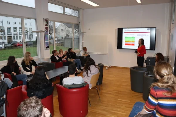 Eine Schülerin steht vor einem Bildschirm und hält vor einer Gruppe Schüler*innen einen Vortrag.