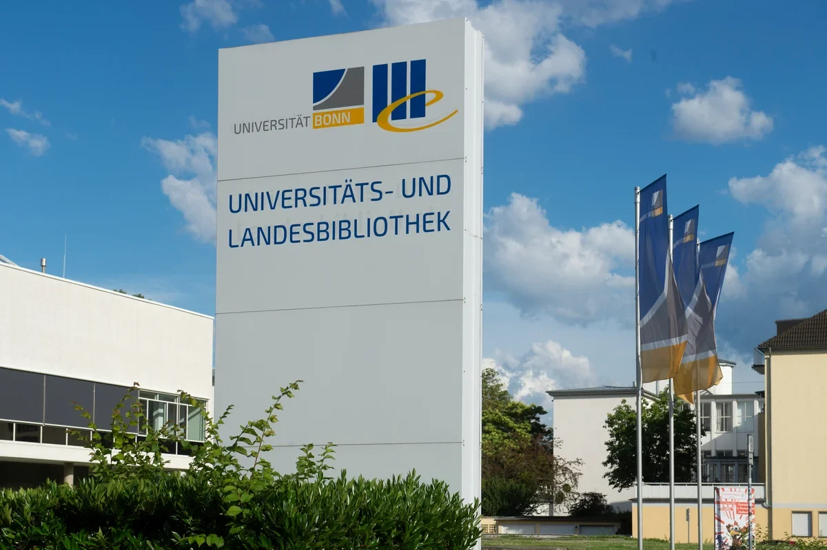 Eine Wissenschaftlerin und ein Wissenschaftler arbeiten hinter einer Glasfassade und mischen Chemikalien mit Großgeräten.