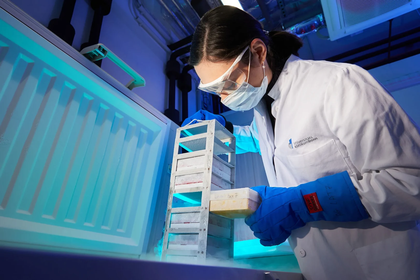 Eine Wissenschaftlerin und ein Wissenschaftler arbeiten hinter einer Glasfassade und mischen Chemikalien mit Großgeräten.